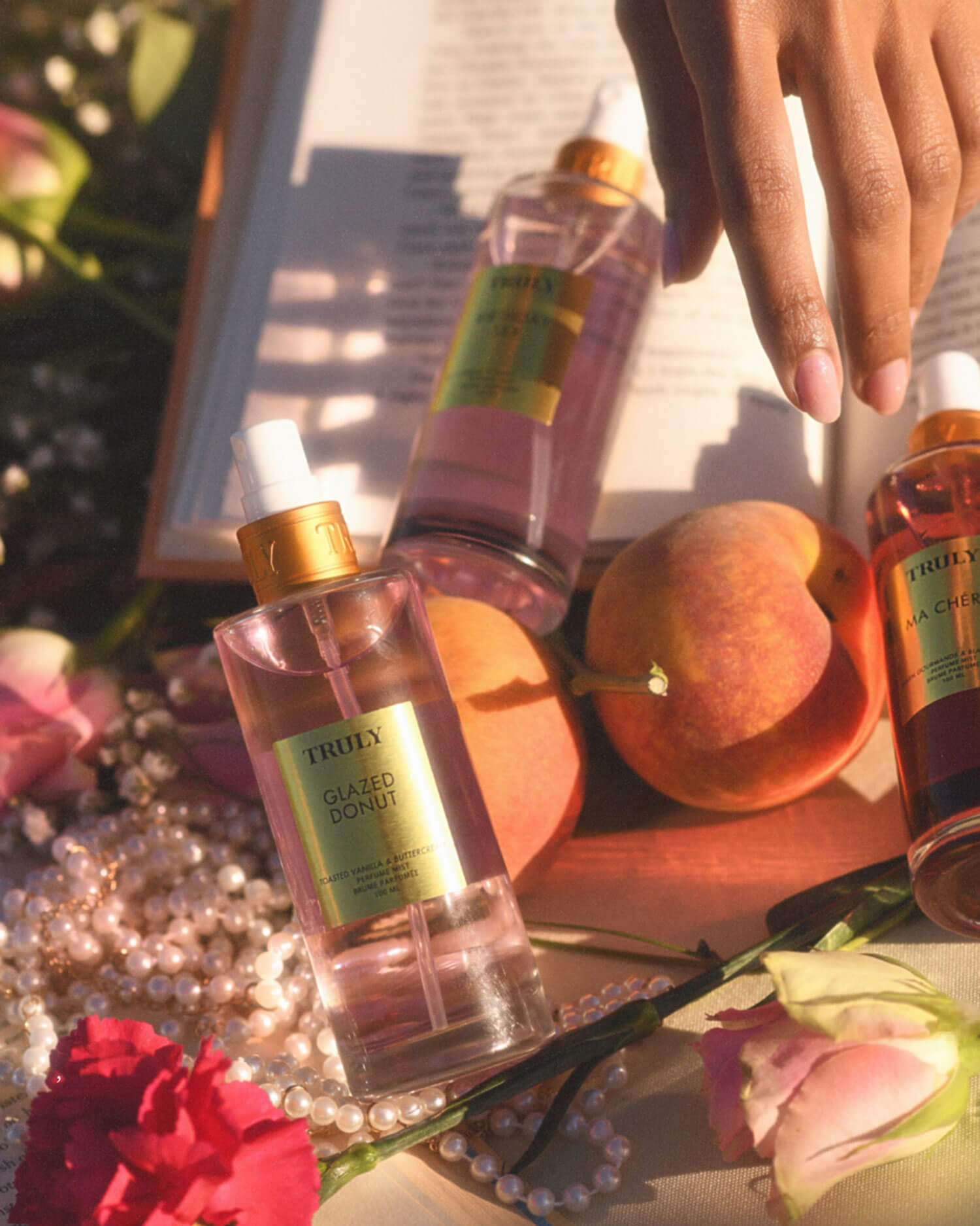 A hand reaches over a book surrounded by Truly Beauty Glazed Donut Perfume Mist bottles, peaches, pink roses, and a pearl necklace, all bathed in warm sunlight with hints of toasted vanilla and whipped buttercream in the air.