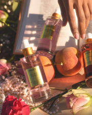 A hand reaches over a book surrounded by Truly Beauty Glazed Donut Perfume Mist bottles, peaches, pink roses, and a pearl necklace, all bathed in warm sunlight with hints of toasted vanilla and whipped buttercream in the air.