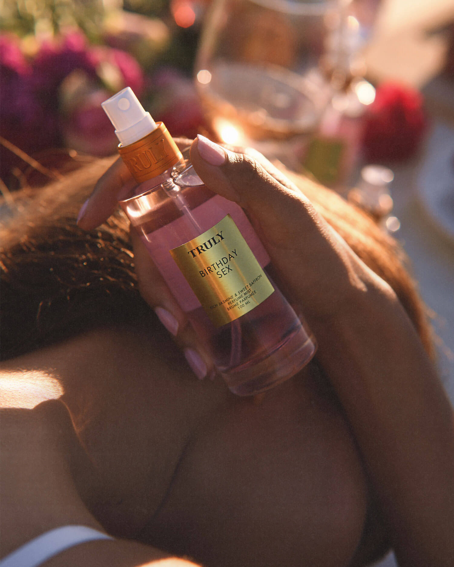 A person with light pink nails holds a bottle of Truly Beauty Birthday Sex Perfume Mist, a spicy floral scent, against a background of blurred flowers and gentle sunlight.
