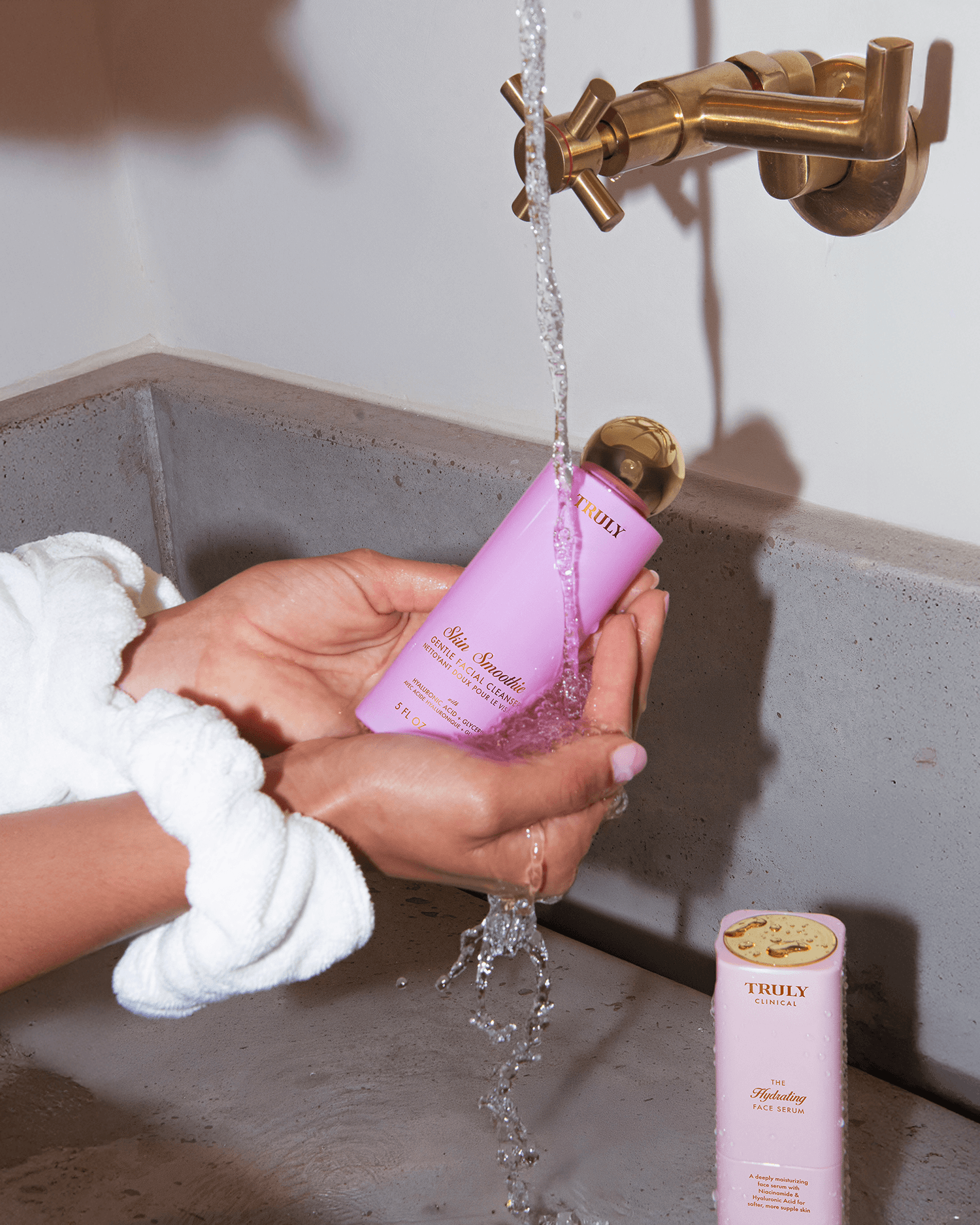 A person in a white robe holds a Truly Beauty Skin Smoothie Gentle Facial Cleanser bottle under running water from a brass faucet, while another identical bottle is placed on the concrete sink.