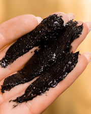 A close-up of a hand with fingers slightly spread, covered in Truly Beauty’s 24K Gold Black Soap Luxury Impurity Cleanser, set against a soft golden-yellow background.
