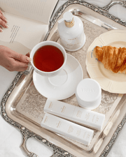 A hand holds a cup of tea on a silver tray with Truly Beauty skincare products, including the Cloud Cream Pro-Peptide Facial Moisturizer, a jar, two labeled tubes, a croissant on a plate, and a partly visible open book nearby.