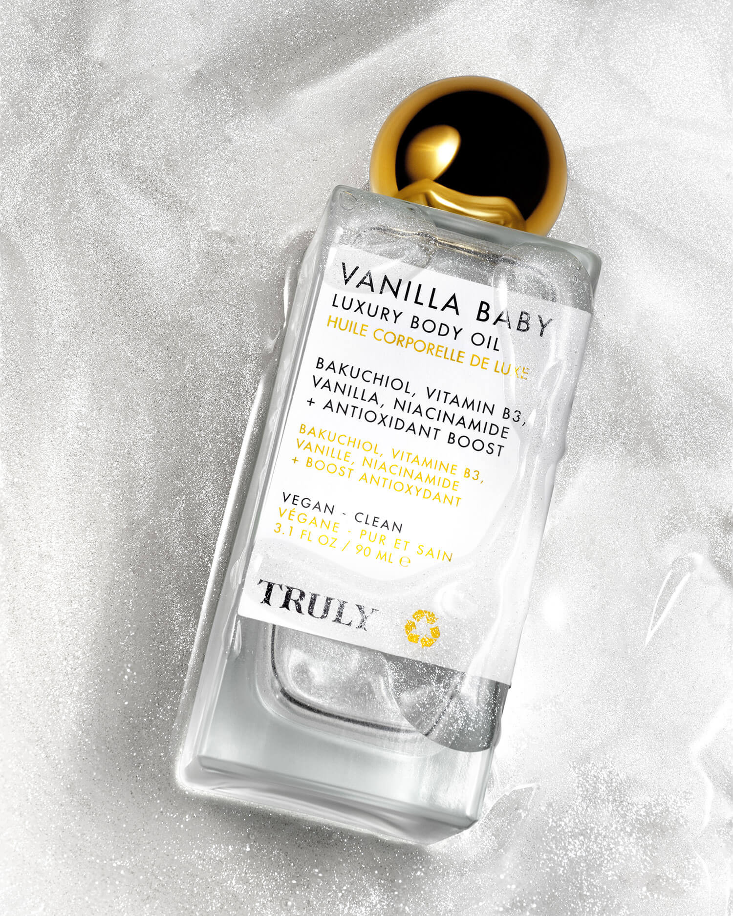 A clear bottle of Truly Beautys Vanilla Baby Luxury Body Oil with a gold cap rests on a white textured surface. The label features plumping bakuchiol, vitamin B3, niacinamide, and notes its vegan and clean.