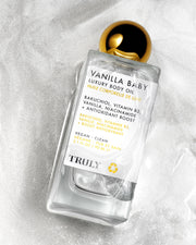 A clear bottle of Truly Beautys Vanilla Baby Luxury Body Oil with a gold cap rests on a white textured surface. The label features plumping bakuchiol, vitamin B3, niacinamide, and notes its vegan and clean.