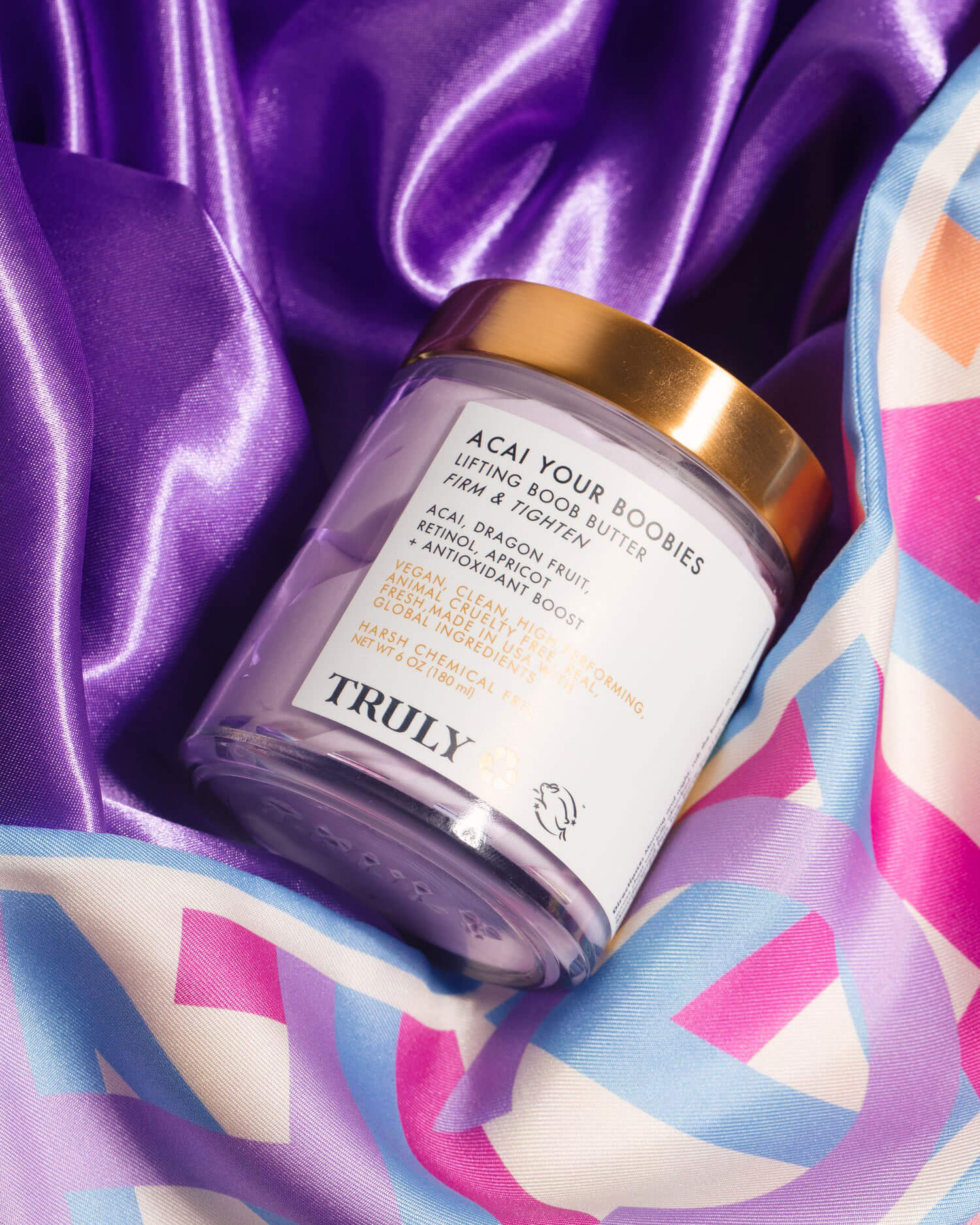 A jar of Truly Beauty’s Acai Your Boobies Butter with a gold lid sits on shiny purple satin, showcasing its whipped formula for tightening and firming, beside a colorful patterned fabric in blue, pink, and orange tones.
