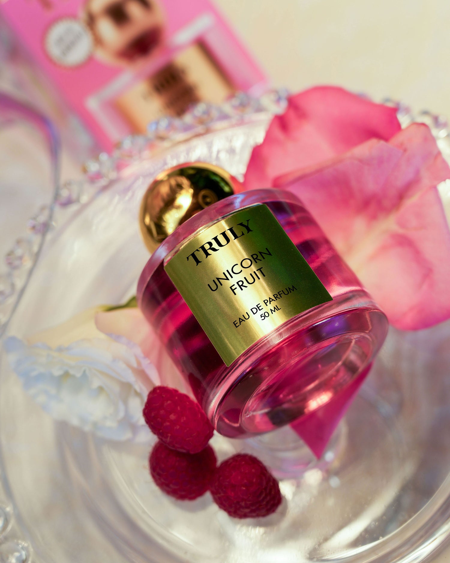 A bottle of Truly Beauty’s Unicorn Fruit Eau De Parfum, with its fruity and feminine scent, rests on a clear glass tray surrounded by pink rose petals, a white flower, raspberries, and the perfume’s pink packaging in the background.