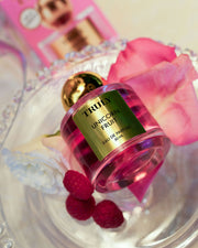 A bottle of Truly Beauty’s Unicorn Fruit Eau De Parfum, with its fruity and feminine scent, rests on a clear glass tray surrounded by pink rose petals, a white flower, raspberries, and the perfume’s pink packaging in the background.