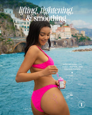 A woman in a bright pink bikini applies Truly Beautys Body Serum Duo by the sea—lifting, tightening & smoothing with watermelon, wheat protein, acai extract & orange flower. A coastal town is visible in the background.