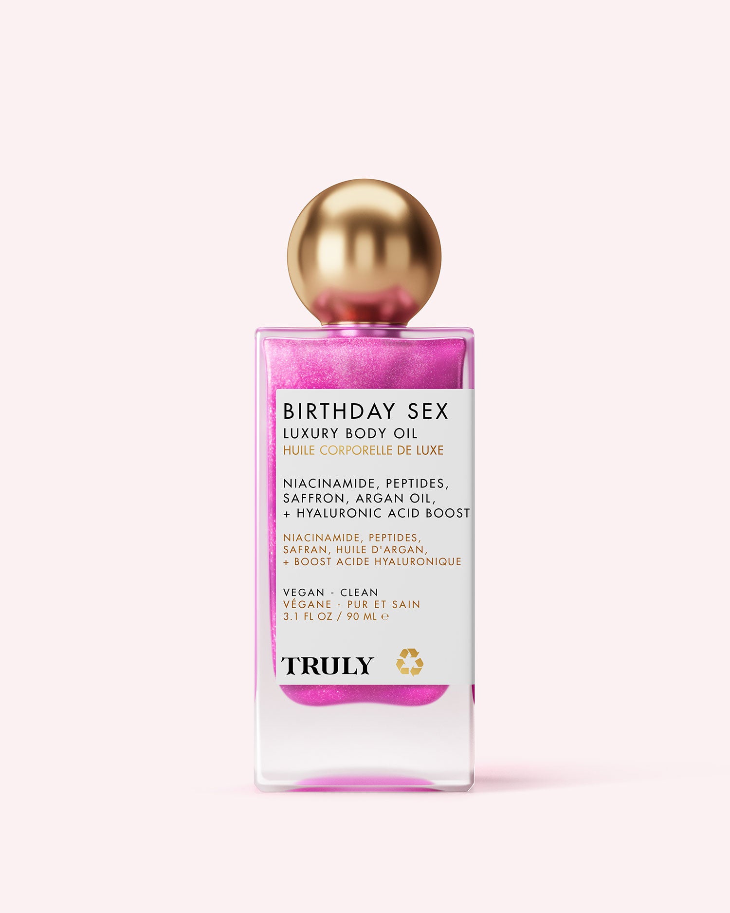 A bottle of Truly Beauty Birthday Sex Luxury Body Oil features pink shimmering liquid, a round gold cap, and a white label listing niacinamide, peptides, hyaluronic acid, saffron, and argan oil.