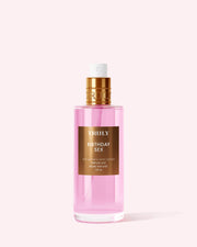 A pink bottle of Truly Beauty’s Birthday Sex Perfume Mist with a gold label and cap sits on a light pink background, offering a spicy floral scent with notes of jasmine and patchouli.