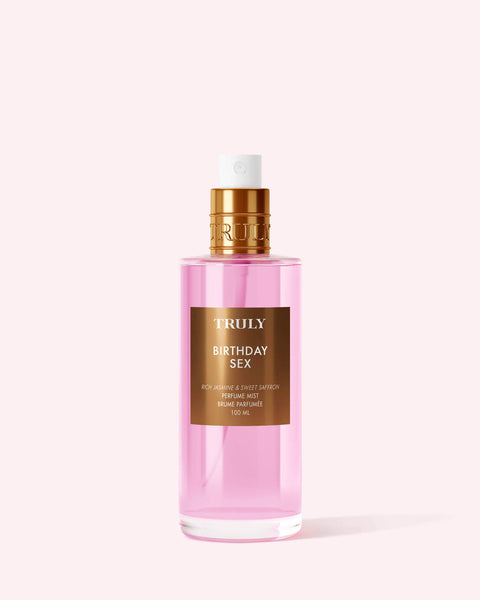 A pink bottle of Truly Beauty’s Birthday Sex Perfume Mist with a gold label and cap sits on a light pink background, offering a spicy floral scent with notes of jasmine and patchouli.