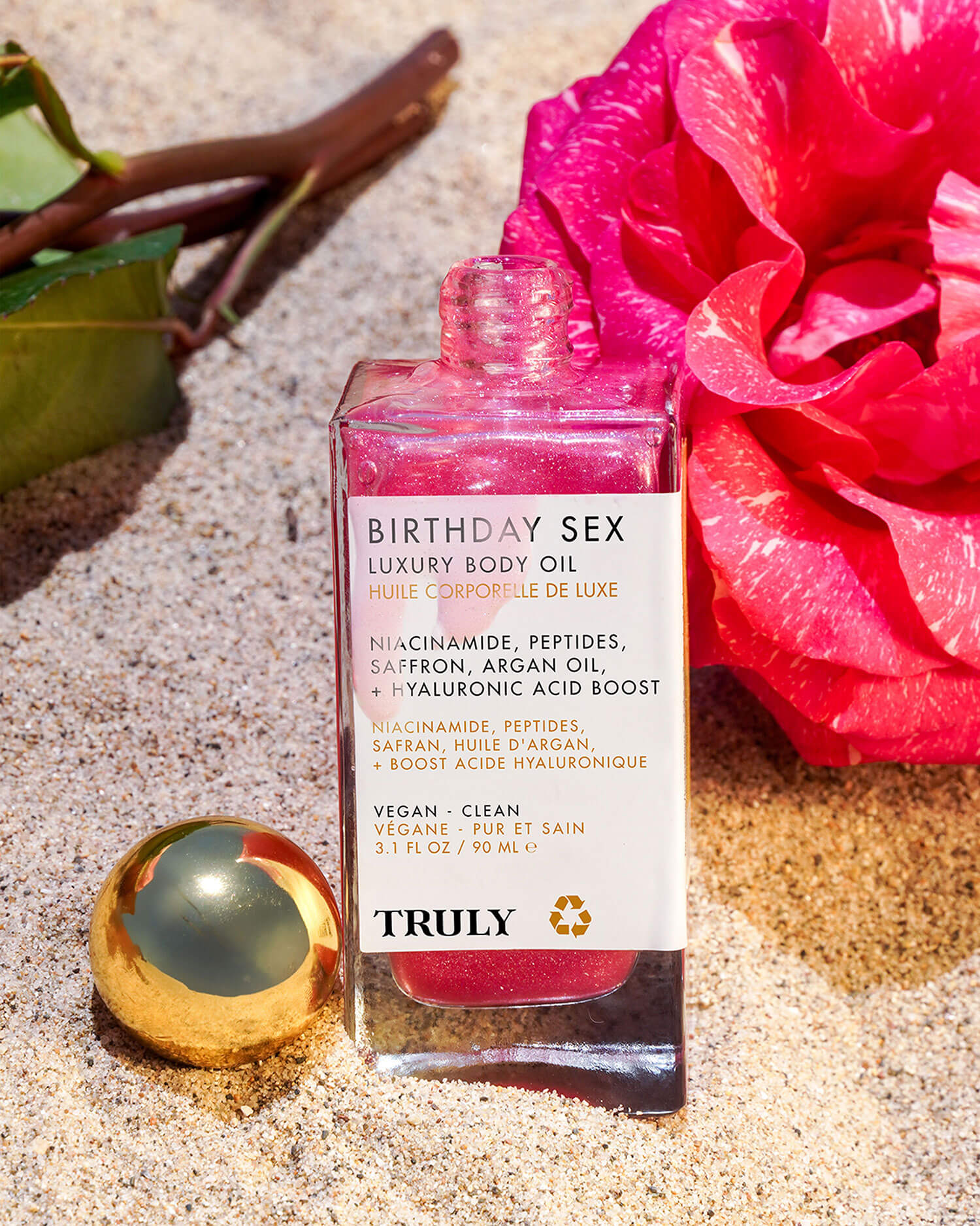 A bottle of Truly Beauty’s Birthday Sex Luxury Trio with a gold cap rests on sand beside a pink rose and green leaves. The shimmering pink oil features jasmine and saffron, with ingredients and product details noted on its label.