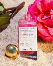 A bottle of Truly Beauty’s Birthday Sex Luxury Trio with a gold cap rests on sand beside a pink rose and green leaves. The shimmering pink oil features jasmine and saffron, with ingredients and product details noted on its label.