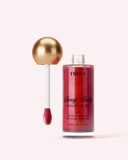 A bottle of Truly Beauty Luxury Peptide Lip Oil with red shimmery oil inside stands beside its gold and white applicator wand on a light pink background.
