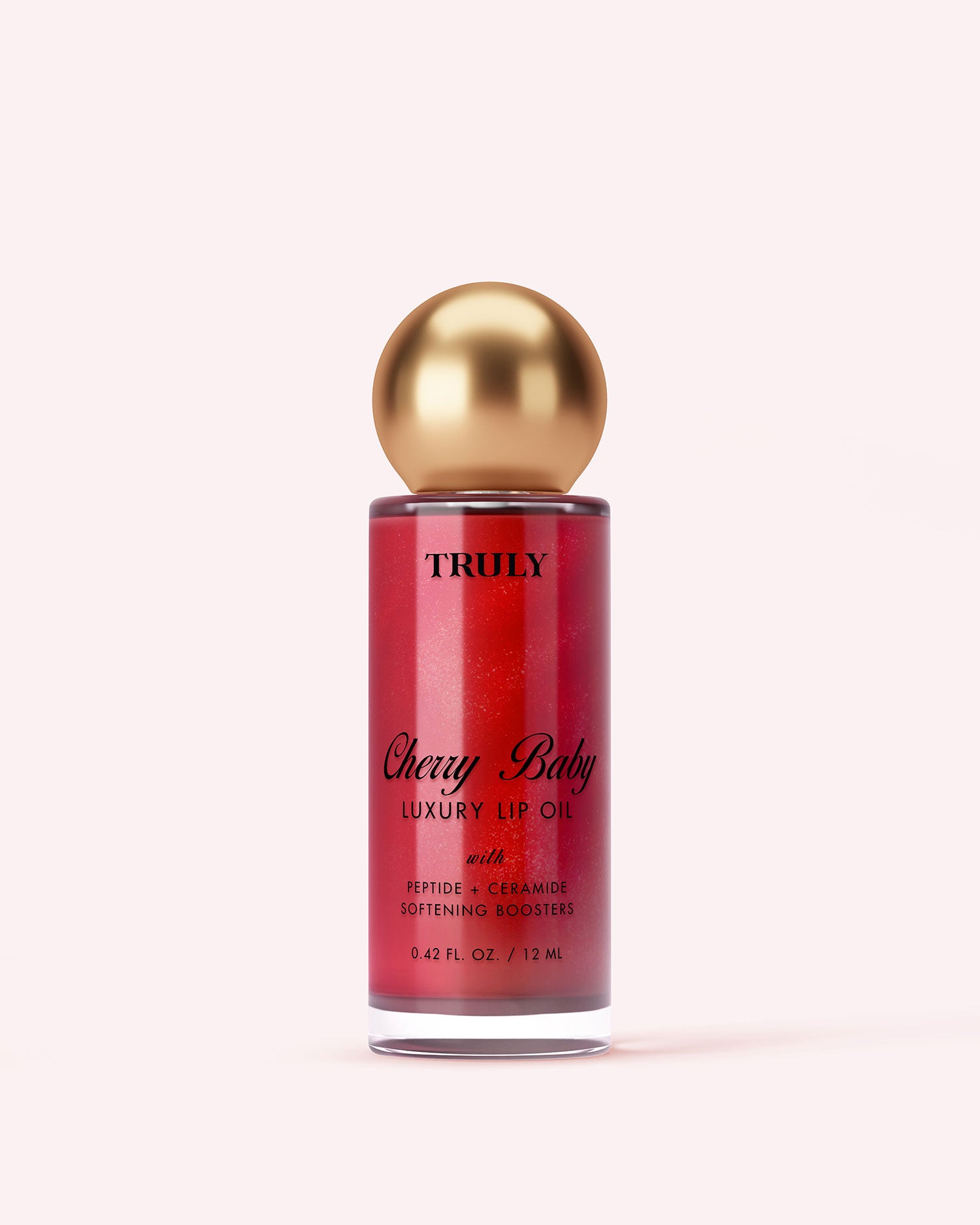 A bottle of Truly Luxury Body Oils by Truly Cherry Baby, with a gold spherical cap and red liquid in a clear container with black text, stands against a pale pink background.