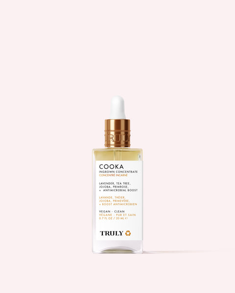 A glass dropper bottle of Truly Beauty’s Cooka Ingrown Concentrate with a gold cap, clear serum, and label listing lavender, tea tree, and jojoba for bikini line care—shown on a pale pink background.