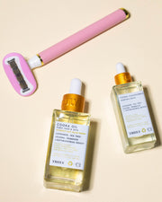 A pink and silver double-bladed razor lies beside two glass dropper bottles from the Cooka Set by Truly Beauty, both containing hair and skin oil for the bikini line, on a beige surface.