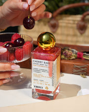 A hand holds a cherry above raspberries next to a bottle of Truly Beauty Cherry Baby Luxury Body Oil with a gold cap, all set on an outdoor table with a wicker basket in the background.