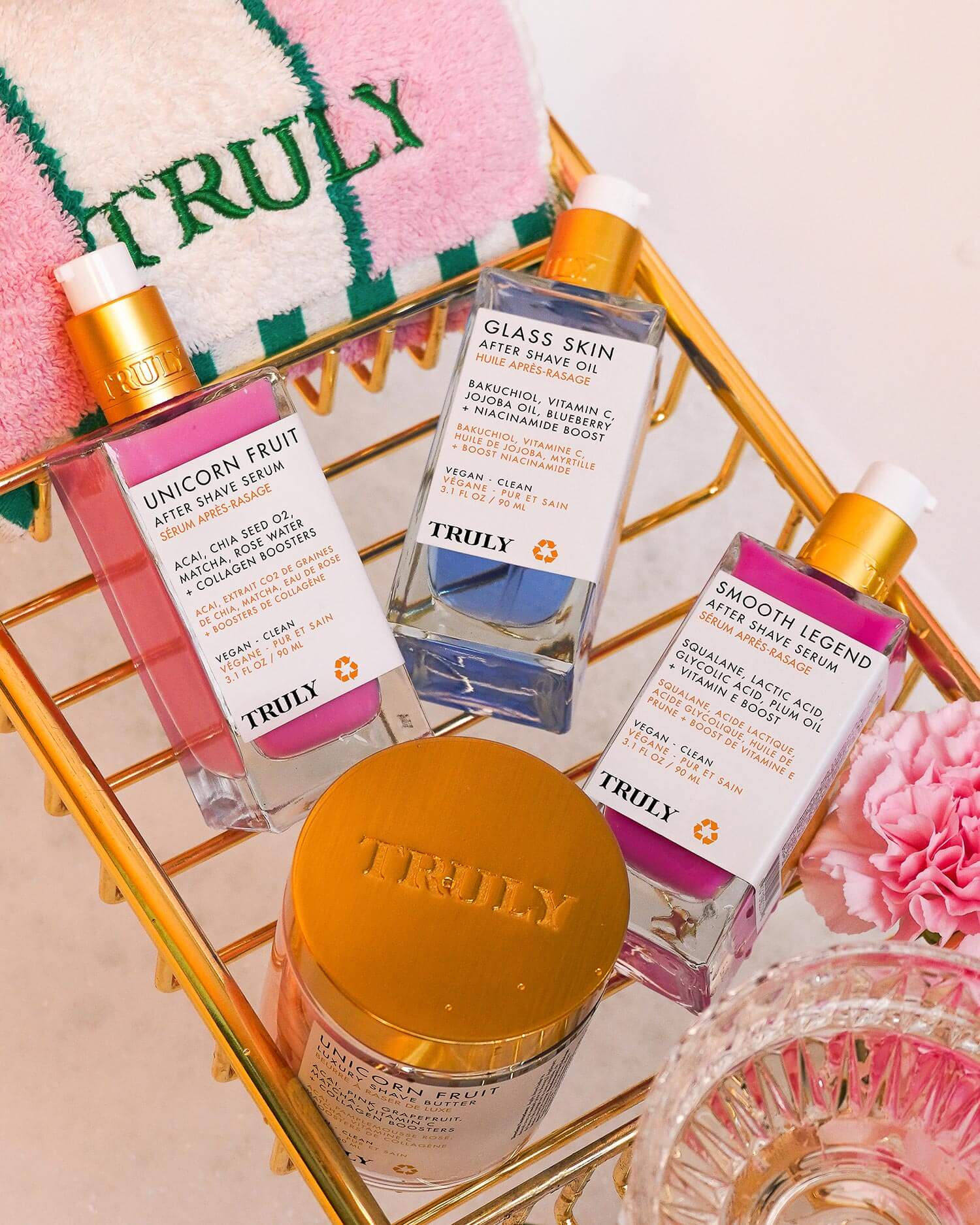 A gold basket holds three Truly Beauty serums, including the Glass Skin After Shave Oil, in pink, blue, and purple liquids with a gold jar. Nearby are a pink Truly Beauty towel and a glass dish—perfect for achieving glass skin.