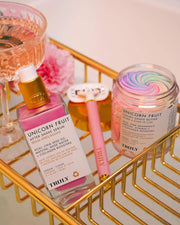 A bottle of Truly Beauty Unicorn Fruit After Shave Serum, a jar of swirled shave butter, and a pink razor sit on a gold wire tray. A pink drink in a glass is nearby.