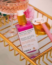 A bottle of Truly Beauty’s Smooth Legend After Shave Serum with exfoliating AHAs and a gold-white pump sits on a gold rack by a glass of pink liquid and a pink razor. The label highlights hydrating, vegan benefits, and key ingredients.