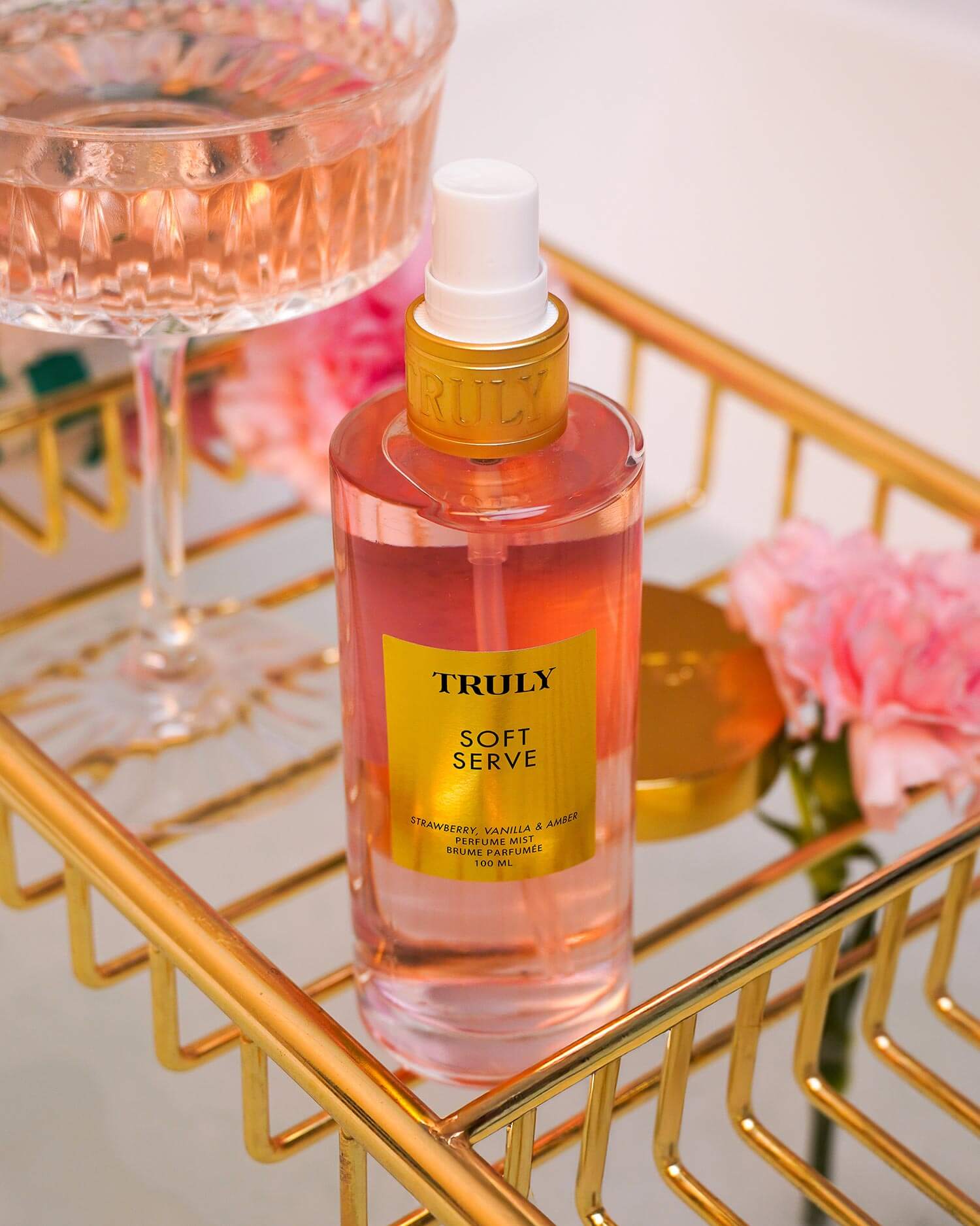 A bottle from the Truly Beauty Soft Serve Luxury Trio with a yellow label rests on a gold tray next to a pink drink and flowers, providing strawberry-scented hydration with peptides and strawberry extract.