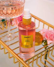 A bottle from the Truly Beauty Soft Serve Luxury Trio with a yellow label rests on a gold tray next to a pink drink and flowers, providing strawberry-scented hydration with peptides and strawberry extract.