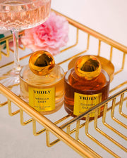Two bottles of Truly Beautys Vanilla Baby Eau De Parfum with gold caps sit in a gold wire basket, evoking notes of amber and caramel drizzle, beside a pink flower and a stemmed glass.