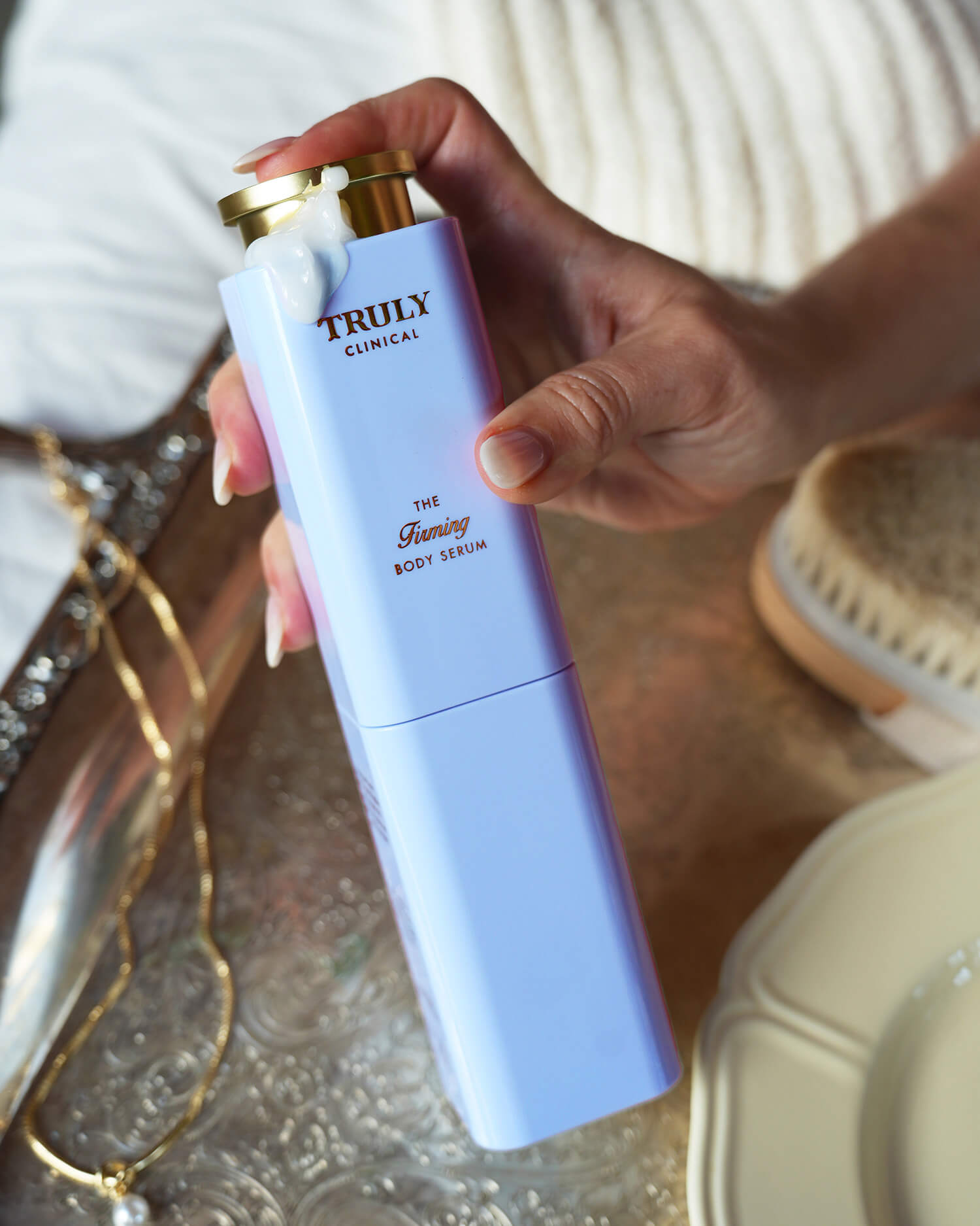 A hand dispenses white lotion from a tall, lavender Truly Beauty bottle labeled The Body Treatments Complete Collection onto its gold cap, with jewelry, a brush, and a plate in the background.