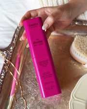 A hand holds a bright pink bottle from Truly Beauty’s The Body Treatments Complete Collection—ideal for targeting stretch marks—over a silver tray, with a body brush and a gold chain nearby.