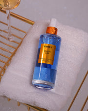 A blue bottle of Clean Girl Perfume Mist by Truly Beauty, featuring a gold cap, sits on a white towel atop a gold rack with part of a wine glass nearby—capturing the fresh floral and clean fragrance vibe.