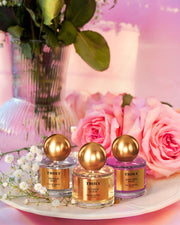 The Perfumers Set by Truly Beauty features three gold-capped perfumes—Vanilla, Golden Gate, and Unicorn—displayed on a pink surface with roses and white flowers, perfect for discovering your signature scent.