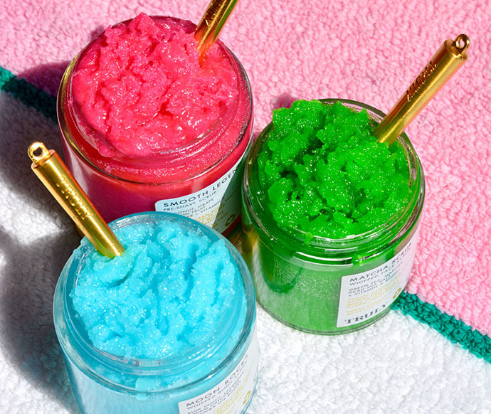Truly’s Mystery Box of 6 Products sits atop a textured white towel with pink and green stripes; three colorful jars, each with a gold spoon, peek out from the set.