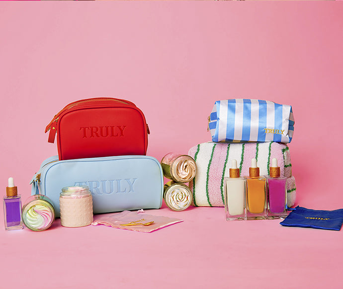 A vibrant assortment of Truly skincare and beauty items from the Truly Mystery Box Of 6 Products, including bags labelled in gold and pastels, bottles, jars, towels, and bath accessories displayed on a pink background.
