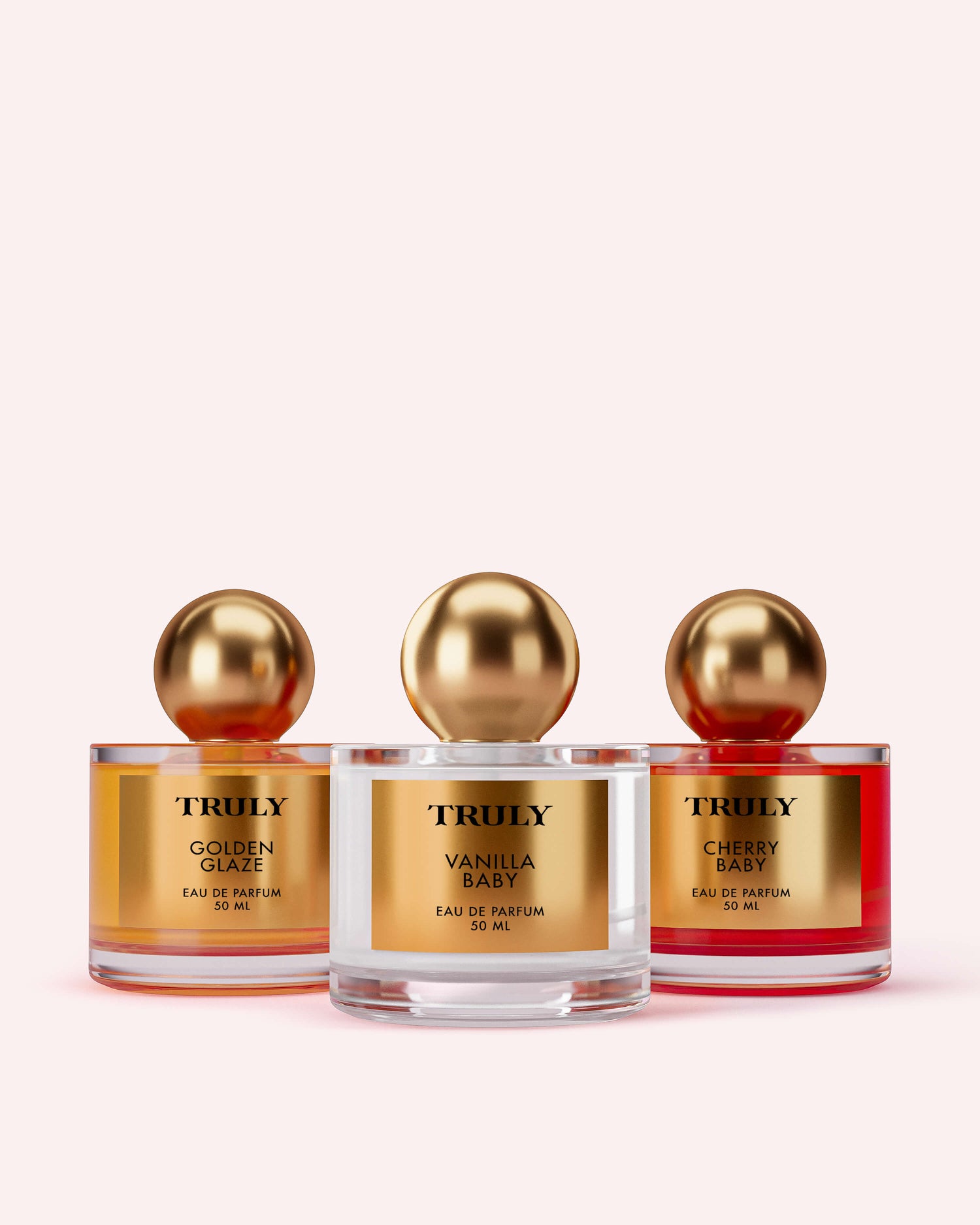 Three glass perfume bottles—Golden Glaze, Vanilla Baby, and Cherry Baby—from the Temptations Collection Eau De Parfum Set by Truly Beauty, shown with gold caps against a light pink background.