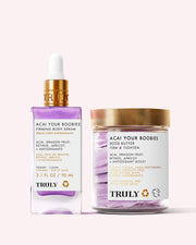 Truly Beauty’s For Your Boobs Routine features a purple-tinted firming serum and a Boob Butter, both in white and gold packaging, designed to firm the chest area. Displayed on a pale pink background.