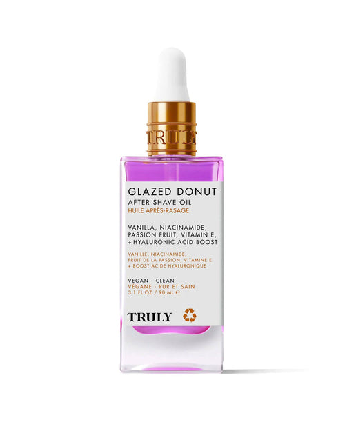 A pink and clear glass dropper bottle labeled Glazed Donut After Shave Oil, highlighting skin-smoothing niacinamide, vitamin E, and hyaluronic acid, is shown on a light pink background.