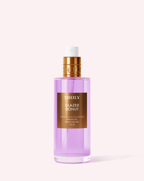 A bottle of Truly Beauty’s Glazed Donut Perfume Mist with a gold cap and pink liquid stands upright on a light pink background, emitting toasted vanilla notes for a dreamy gourmand fragrance experience.