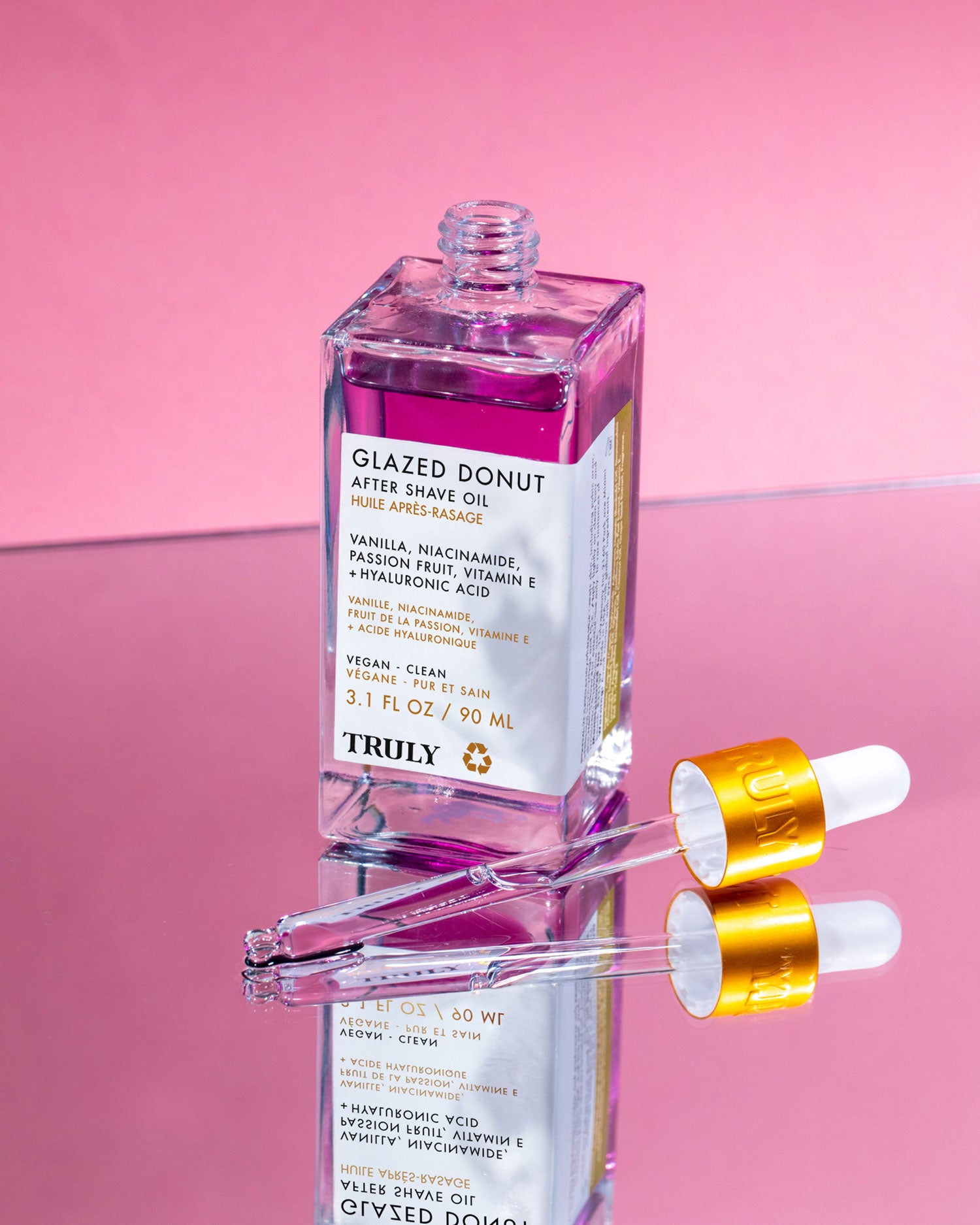 A clear bottle of Truly Beauty Glazed Donut After Shave Oil filled with purple liquid sits on a reflective surface, flanked by two glass droppers with gold caps. The pink background highlights this hydrating daily shave serum.