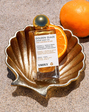 A bottle of Truly Beauty Golden Glaze Luxury Body Oil rests in a golden shell dish on sand, with oranges beside it, creating a beachy and luxurious vibe.