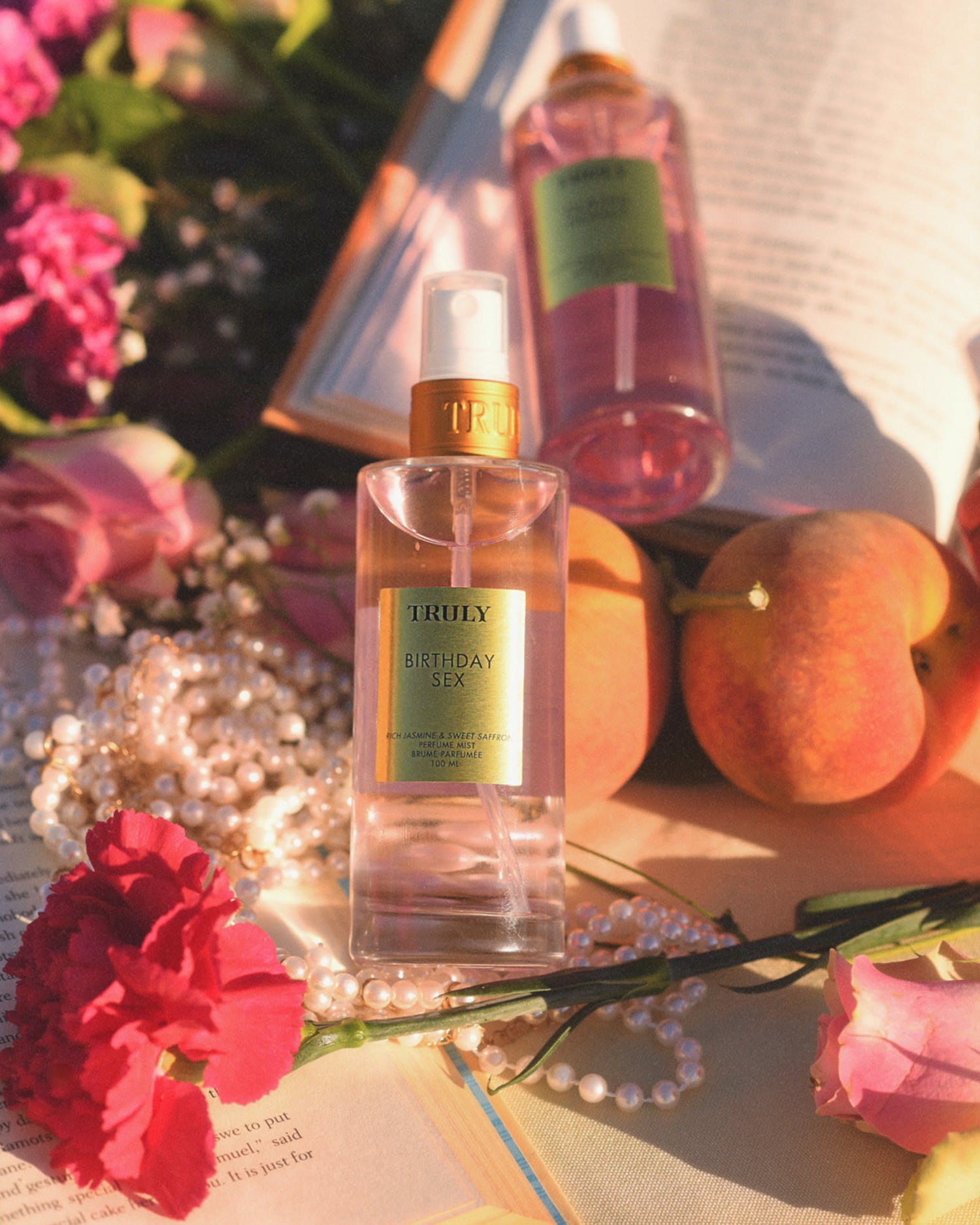 A bottle of Truly Beauty Birthday Sex Perfume Mist rests on an open book amid pink flowers, peaches, pearls, and another pink bottle in warm sunlight—a spicy floral scent with jasmine and patchouli fills the air.