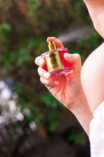 A person sprays Truly Beauty’s Cherry Baby Eau De Parfum on their neck, savoring its long-lasting scent with a blurred green outdoor background.