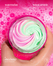 A hand holds an open jar of Truly Beauty’s Buns of Glowry Tighten & Glow Smoothing Butt Butter with pink and green swirls, set against a pink bubbly background. Key ingredients: watermelon extract (plumps), wheat protein (firms), shea butter (hydrates).