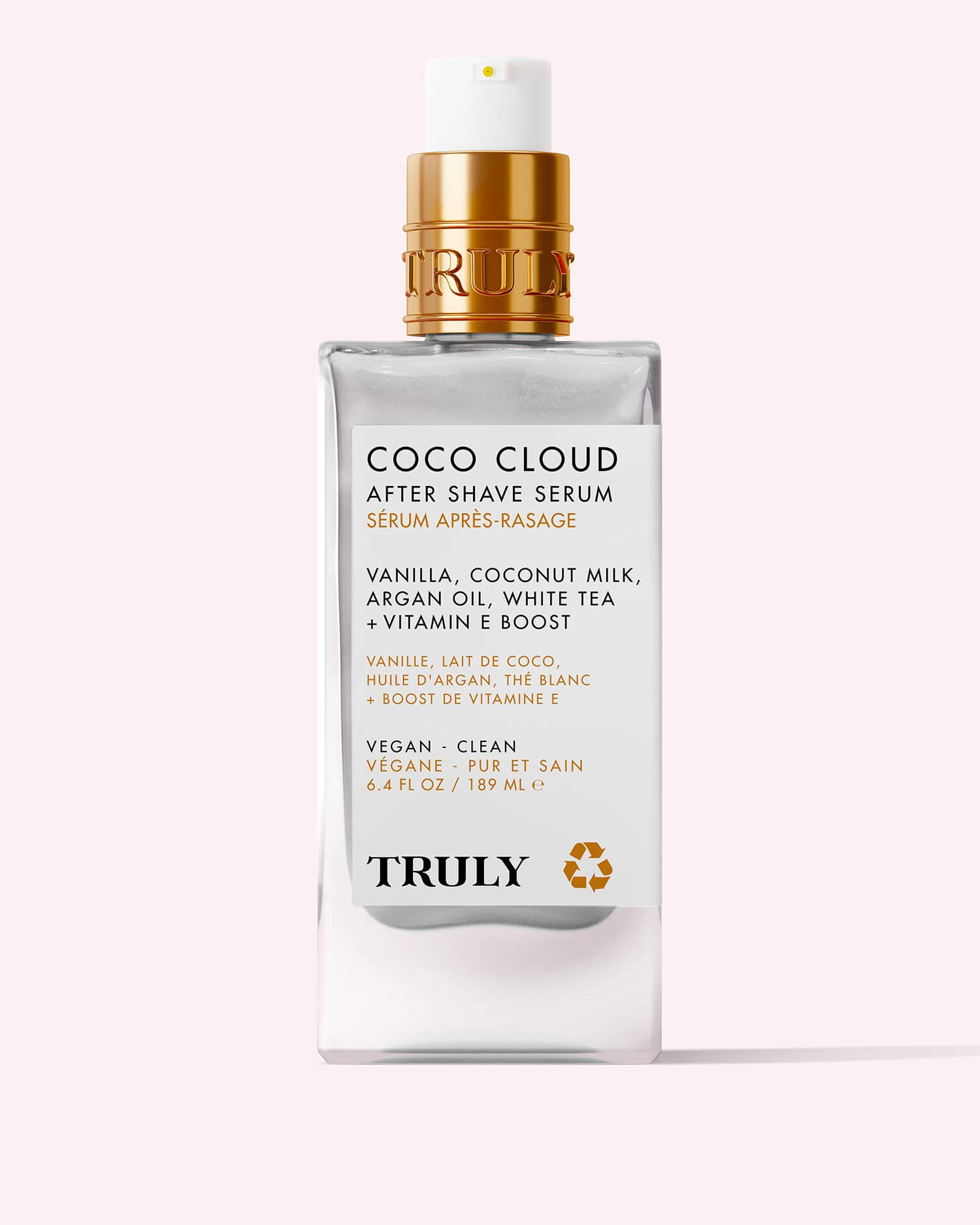 A clear bottle of Truly Beauty’s Coco Cloud After Shave Serum with a gold pump and label featuring coconut, argan oil, and vanilla ingredients, displayed on a light pink background.