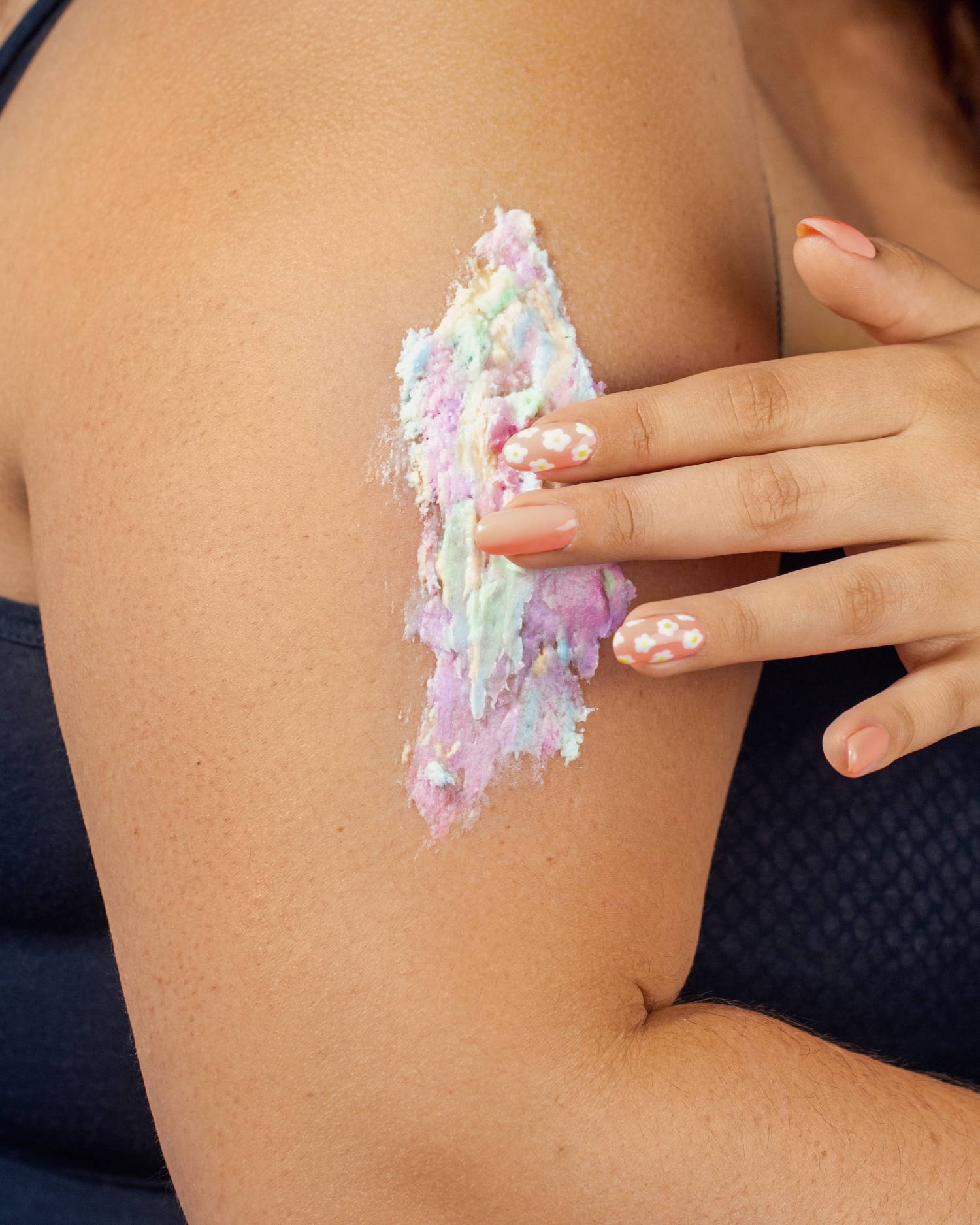 A person applies Truly Beauty’s pastel-colored Unicorn Fruit Duo body butter with a cotton candy scent to their upper arm. Their peach nails have white flower designs, and they’re wearing a dark, textured sleeveless top.