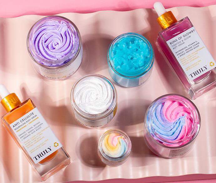 A selection of Truly skincare in pastel jars and gold-capped dropper bottles, labeled “Truly,” arranged on a light pink wavy surface. Features the Truly Mystery Box Of 6 Products.