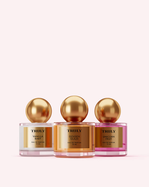The Perfumers Set by Truly Beauty features three perfume bottles with gold spherical caps—Vanilla Baby, Golden Glaze, and Unicorn Fruit—lined up on a light pink background, offering an irresistible fragrance discovery.