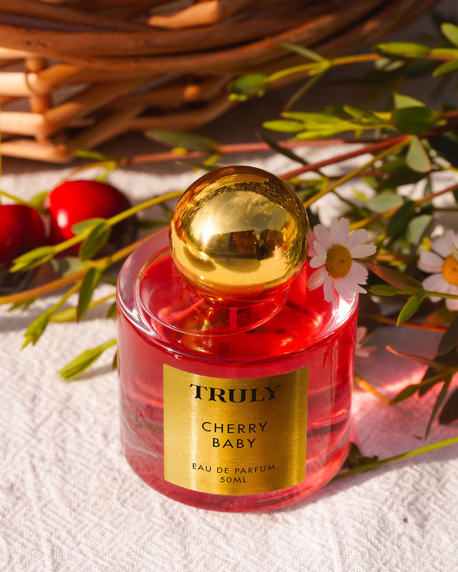 A pink glass bottle from the Truly Beauty Temptations Collection Eau De Parfum Set rests on a white cloth with daisies, green stems, and cherries nearby, framed by a wicker basket in the background.