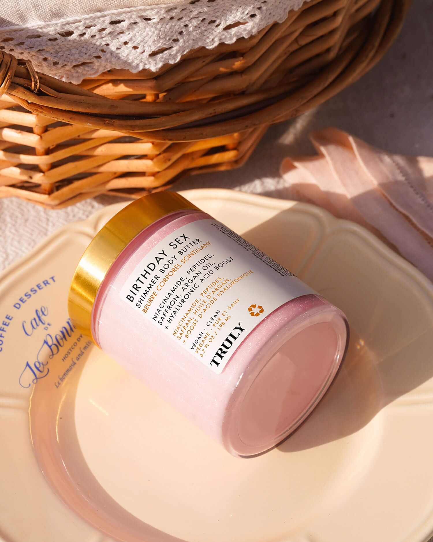 A jar of Truly Beauty Birthday Sex Shimmer Body Butter rests on a cream plate beside a woven basket and light pink fabric, glowing in natural sunlight.