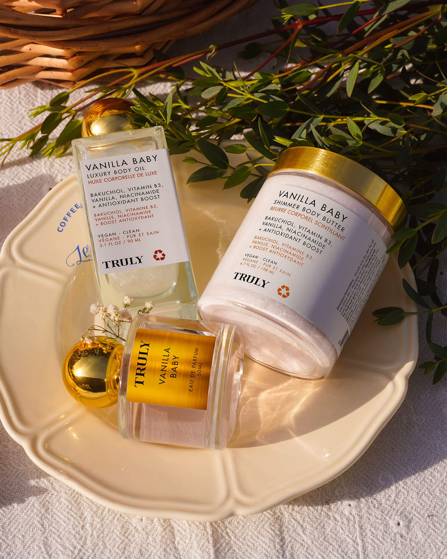 The Vanilla Baby Luxury Trio by Truly Beauty—a body oil, body butter, and shimmer polish—rests on a cream plate with greenery and sunlight, capturing a warm vanilla scent and the promise of glowing skin.